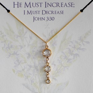 May include: A gold necklace with a pendant featuring three clear crystal stones. The pendant is a drop design with the largest stone at the top and the smallest stone at the bottom. The necklace is on a white card with the text "He must increase; I must decrease John 3:30".