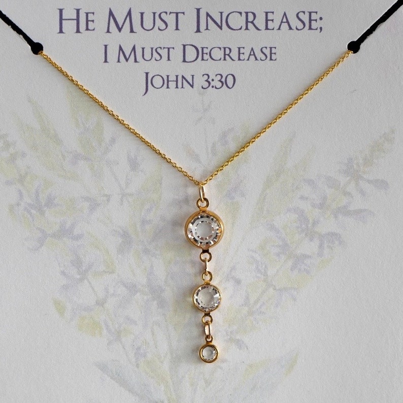 May include: A gold necklace with a pendant featuring three clear crystal stones. The pendant is a drop design with the largest stone at the top and the smallest stone at the bottom. The necklace is on a white card with the text "He must increase; I must decrease John 3:30".