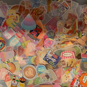 May include: A collection of colorful stickers featuring various designs, including fruit, animals, and text. The stickers are arranged in a random pattern on a white surface.