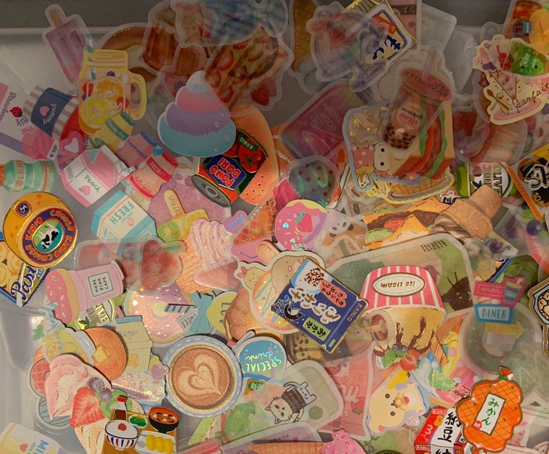 May include: A collection of colorful stickers featuring various designs, including fruit, animals, and text. The stickers are arranged in a random pattern on a white surface.