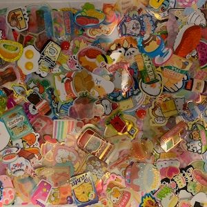 May include: A large collection of colorful and whimsical stickers featuring cartoon characters, food, and other cute designs. The stickers are arranged in a random pattern and are perfect for decorating journals, laptops, or other personal items.