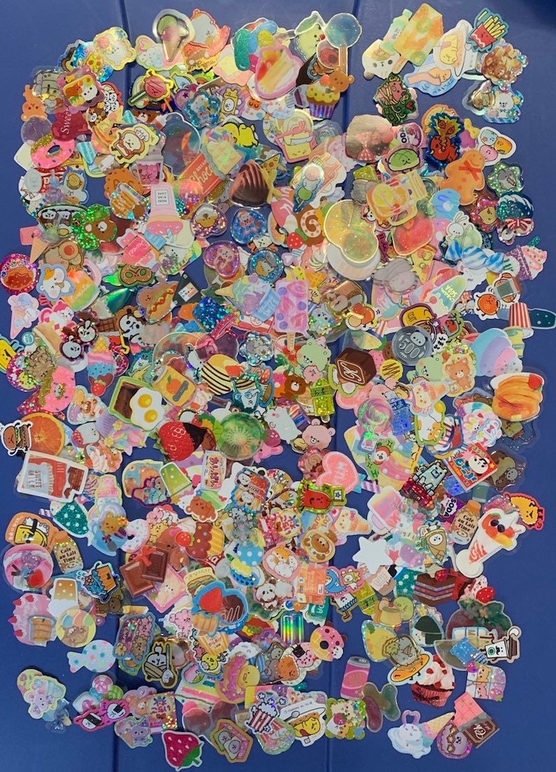 May include: A large collection of colorful, sparkly, and holographic stickers featuring various cute cartoon characters, food, and other designs. The stickers are arranged in a random pattern on a blue surface.