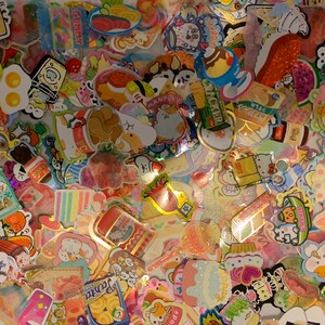 May include: A collection of colorful, glittery stickers featuring various cartoon characters, food, and other designs. The stickers are arranged in a random pattern and are perfect for decorating journals, planners, and other items.