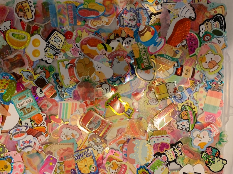 May include: A collection of colorful, glittery stickers featuring various cartoon characters, food, and other designs. The stickers are arranged in a random pattern and are perfect for decorating journals, planners, and other items.