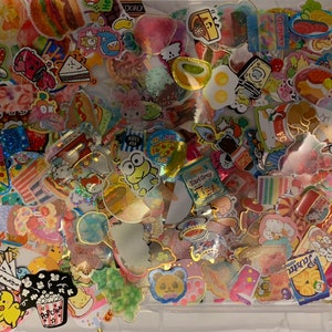 May include: A clear plastic container filled with colorful stickers featuring various food and cartoon characters. The stickers are mostly in shades of pink, blue, yellow, and green. Some of the stickers have glitter accents. The stickers are arranged in a random pattern.