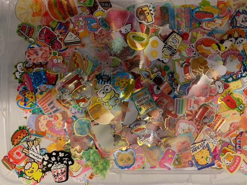 May include: A clear plastic container filled with colorful stickers featuring various food and cartoon characters. The stickers are mostly in shades of pink, blue, yellow, and green. Some of the stickers have glitter accents. The stickers are arranged in a random pattern.
