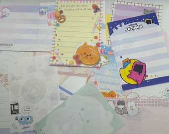 May include: A collection of colorful stationery featuring cute animal illustrations. The stationery includes lined paper, notepads, and memo pads. The designs include a cat with a pancake, a pink monster, a blue monster, and a hamburger.