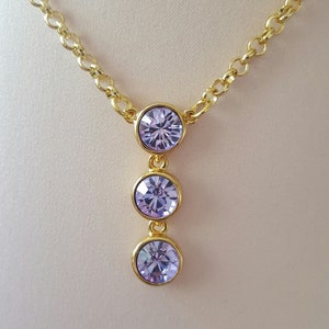 Puede incluir: Un collar de cadena dorada con un colgante que presenta tres piedras redondas de color púrpura claro.