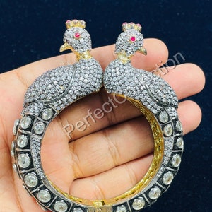 May include: A gold bangle bracelet with two peacock designs encrusted with clear and red gemstones. The bracelet is hinged and has a clasp.