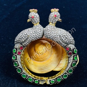 May include: A gold bangle bracelet with two peacocks facing each other. The peacocks are adorned with clear and red gemstones. The bangle is decorated with green and black gemstones.