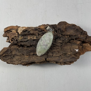 Może przedstawiać: Owalny wisiorek ze srebrnym uchwytem. Wisiorek posiada jasnozielony kamień z fioletowymi żyłkami, osadzony w srebrnej metalowej ramie. Wisiorek jest prezentowany na kawałku teksturowanego, ciemnobrązowego drewna.