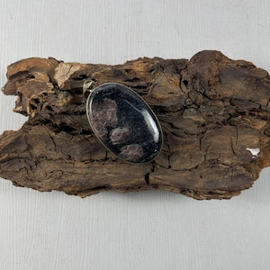 Peut inclure: Pendentif ovale avec une surface sombre et mouchetée et une bordure en métal argenté. Le pendentif a une finition polie et brillante et est présenté sur un morceau de bois brun texturé.