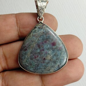 May include: A teardrop-shaped pendant featuring a polished, multi-coloured stone with mottled grey, blue, and deep pink inclusions. The stone is set in a smooth silver-toned metal bezel with an ornate, engraved silver-toned triangular bail.