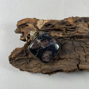 Puede incluir: Un colgante cuadrado con una piedra oscura y moteada engastada en un marco de metal plateado. La piedra tiene una base negra con inclusiones granates y rojo oscuro. El colgante se muestra sobre un trozo de madera marrón oscuro.
