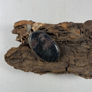 以下が含まれることがあります： 銀色の金属フレームにセットされた、暗く斑点のある石の楕円形のペンダント。石は黒と栗色の色合いが混ざっています。ペンダントは、質感のある濃い茶色の木片の上に展示されています。