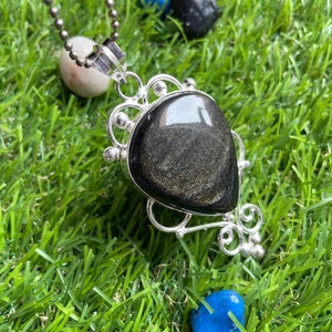 May include: A silver chain necklace with a black and gold gemstone pendant. The pendant is set in a silver filigree design. The necklace is photographed on a green surface.