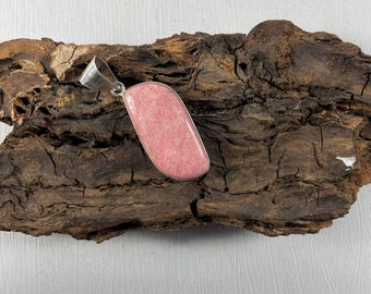 May include: A pink gemstone pendant with a silver bail. The pendant is rectangular with rounded edges. The gemstone has a speckled appearance. The pendant is displayed on a piece of dark brown, textured wood.