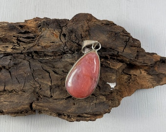 May include: A teardrop-shaped pendant with a silver-colored bail. The pendant features a polished, pink gemstone with a silver-colored bezel. The pendant is displayed on a piece of dark brown, textured wood.