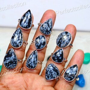 Puede incluir: Una colección de diez anillos de plata con engastes de piedra preciosa de obsidiana copo de nieve. Los anillos se muestran en una mano, cada anillo presenta una piedra de obsidiana copo de nieve diferente.
