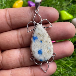 Può includere: Un ciondolo d'argento con una pietra blu e bianca a forma di lacrima. La pietra ha un motivo maculato ed è incastonata in una cornice d'argento con intricati disegni floreali.