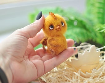 Può includere: Un piccolo pulcino di feltro giallo con occhi neri e un becco, seduto in una mano. Il pulcino è in un nido fatto di paglia e ha un guscio d'uovo rotto accanto.