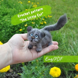 Puede incluir: Un gato gris tejido a ganchillo con ojos azules y una cola larga. El gato se sostiene en una mano, con el texto "Crochet pattern 'The Little Cat'" en una pancarta verde. También es visible el texto "Eng/PDF".