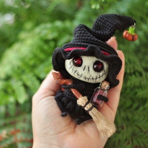 May include: A small crocheted witch doll with a black hat, red trim, and a stitched-on face. The doll is holding a small broom and a bottle of potion.