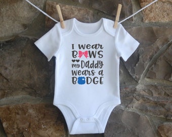 Puede incluir: Un body blanco para bebé con el texto "I wear BOWS my Daddy wears a BADGE" con un lazo rosa y una insignia azul.