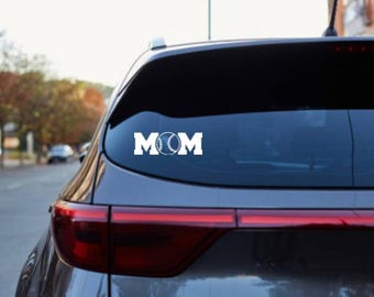 Puede incluir: Calcomanía de coche de vinilo blanco con la palabra "MOM" y un gráfico de béisbol. La calcomanía está aplicada en la ventana trasera de un coche gris. La imagen está tomada al aire libre con un fondo borroso de árboles y una calle.