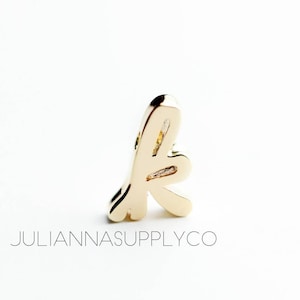May include: Gold charm in the shape of the letter "K" with a stylised, abstract design.