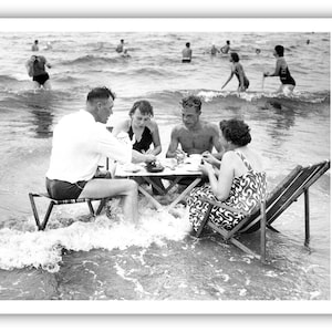 Puede incluir: Fotograf&iacute;a en blanco y negro de un grupo de personas haciendo un picnic en el oc&eacute;ano. Est&aacute;n sentados alrededor de una peque&ntilde;a mesa, con las olas a su alrededor. Otras personas nadan en el fondo.