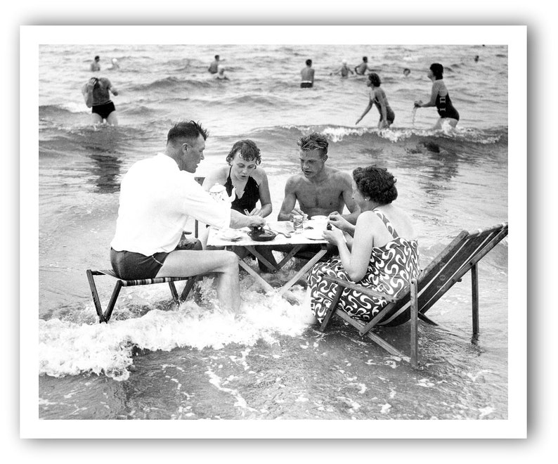 Puede incluir: Fotograf&iacute;a en blanco y negro de un grupo de personas haciendo un picnic en el oc&eacute;ano. Est&aacute;n sentados alrededor de una peque&ntilde;a mesa, con las olas a su alrededor. Otras personas nadan en el fondo.
