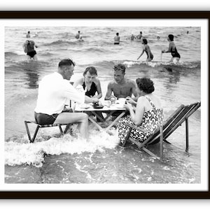 Puede incluir: Fotograf&iacute;a en blanco y negro de un grupo de personas comiendo en una mesa instalada en el oc&eacute;ano. La mesa y las sillas est&aacute;n parcialmente sumergidas en el agua. Otras personas nadan y chapotean en el fondo.
