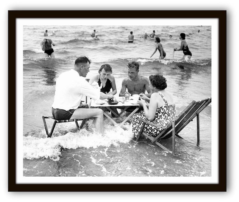 Puede incluir: Fotograf&iacute;a en blanco y negro de un grupo de personas comiendo en una mesa instalada en el oc&eacute;ano. La mesa y las sillas est&aacute;n parcialmente sumergidas en el agua. Otras personas nadan y chapotean en el fondo.