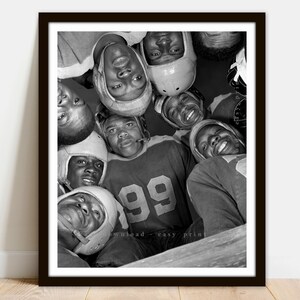 May include: Black and white photo of a group of young men wearing football uniforms and helmets. They are huddled together, looking up at the camera. The player in the center is wearing a jersey with the number 99.