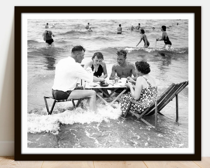 Puede incluir: Impresi&oacute;n enmarcada en blanco y negro de una escena de playa vintage. Un grupo de personas est&aacute; sentado alrededor de una peque&ntilde;a mesa en el agua, comiendo. Otras personas nadan en el fondo. La impresi&oacute;n est&aacute; en un marco negro.