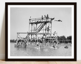 May include: Black and white vintage photograph of a wooden diving platform on a lake. People in swimsuits are on the platform, some diving into the water. A few people are in a small boat.