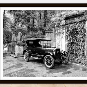May include: A black and white photograph of a vintage Ford Model T touring car parked in front of a stone wall covered in ivy. The car is in good condition and has a classic look.