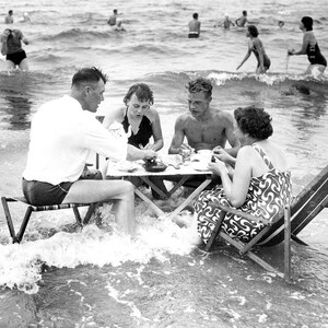 Puede incluir: Fotograf&iacute;a en blanco y negro de un grupo de personas haciendo un picnic en el oc&eacute;ano. Est&aacute;n sentados en una mesa peque&ntilde;a y en sillas plegables, con las olas rompiendo suavemente a su alrededor. Otras personas nadan en el fondo.