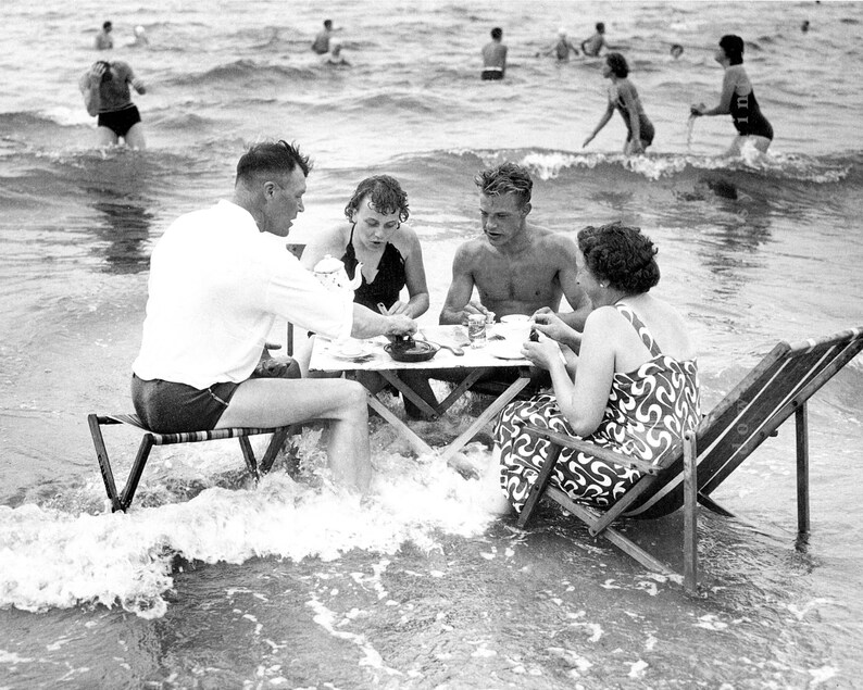 Puede incluir: Fotograf&iacute;a en blanco y negro de un grupo de personas haciendo un picnic en el oc&eacute;ano. Est&aacute;n sentados en una mesa peque&ntilde;a y en sillas plegables, con las olas rompiendo suavemente a su alrededor. Otras personas nadan en el fondo.