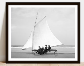 May include: Black and white photograph of four people riding a land yacht on a beach. The land yacht has a large sail and is being pulled by the wind.