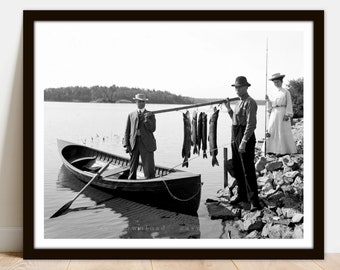 May include: A black and white photograph of three people fishing. Two people stand on a rocky shore, while one person stands in a wooden boat. The people are holding fishing rods and a string of fish.