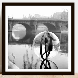 May include: A black and white photograph of a woman standing inside a large clear bubble. The woman is wearing a dress and a scarf. The bubble is floating on the water in front of a bridge. The bridge is in the background of the image.