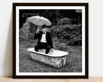 May include: A man in a tuxedo and a bow tie sits in a white bathtub holding a red and white umbrella. The bathtub is outdoors on a grassy lawn.