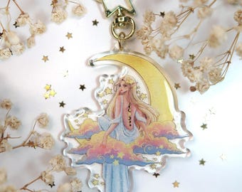 Può includere: Un portachiavi a tema celeste con una figura seduta con lunghi capelli biondi, ornata da una luna crescente e stelle. Il design include una chiusura a forma di stella color oro e un anello color oro. La figura è seduta su una nuvola dai colori pastello.