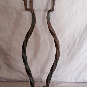 Peut inclure: Objet décoratif en fer forgé. Il présente deux supports torsadés et incurvés, reliés par des rouleaux métalliques. L'ensemble a un aspect rustique et vieilli, avec des marques de soudure visibles et une finition sombre et patinée.