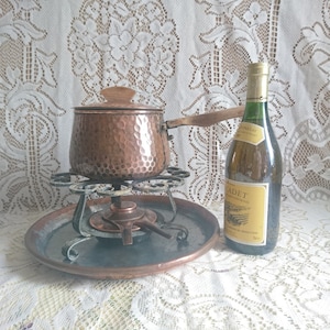 Peut inclure: Un pot à fondue en cuivre avec un couvercle et une poignée en bois posé sur un plateau en cuivre avec un support en métal. Une bouteille de vin blanc avec une étiquette qui indique "Cadet" est à côté du pot.