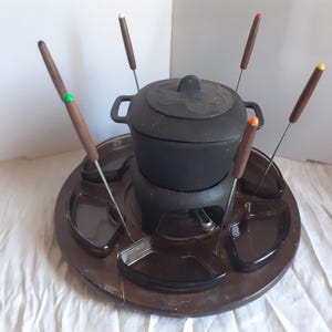 May include: A black fondue pot with a matching lid, set on a brown serving tray with individual compartments. Includes six fondue forks with wooden handles and colored tips. A complete set for entertaining.