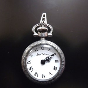 Puede incluir: Un reloj de bolsillo plateado con una esfera blanca y marcadores de hora romanos negros. El reloj tiene una cadena plateada decorativa unida a la parte superior y las palabras "Jean Palice" impresas en la esfera.