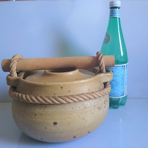 Peut inclure: Un pot en céramique brun avec une poignée en bois et une corde attachée autour du haut. Le pot a un glaçage moucheté et repose sur une surface blanche. Une bouteille d'eau en verre transparent se trouve à l'arrière-plan.